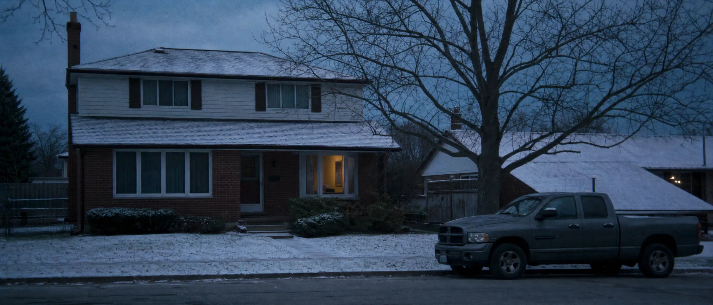 A two-storey house on a snowy suburban street at dusk, one window lit